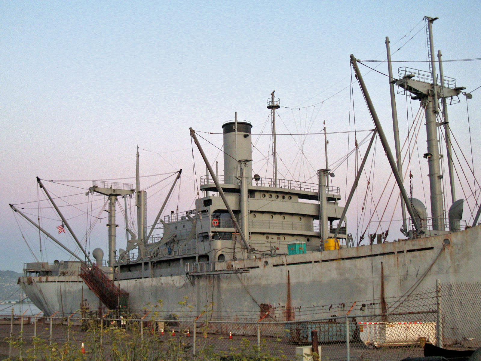 The weathered SS Red Oak Victory docked at the Port of Richmond, seen across a fenced shipyard lot